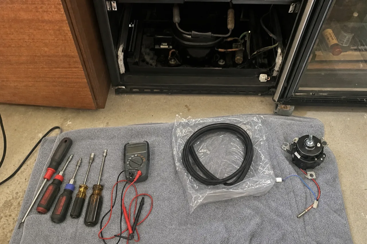 An undercounter refrigeration cabinet pulled out from a timber cabinet run with its lower service panel removed, compressor and tubing visible, tools and a replacement gasket laid out on a towel in front of it.