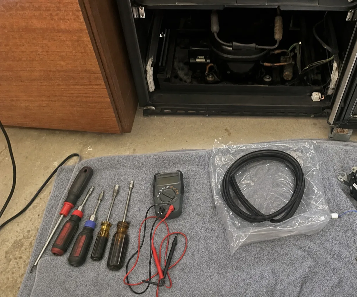 The open base of an undercounter refrigeration cabinet with its rear panel off: black compressor, copper suction line and wiring exposed, hand tools on a towel on the floor below.