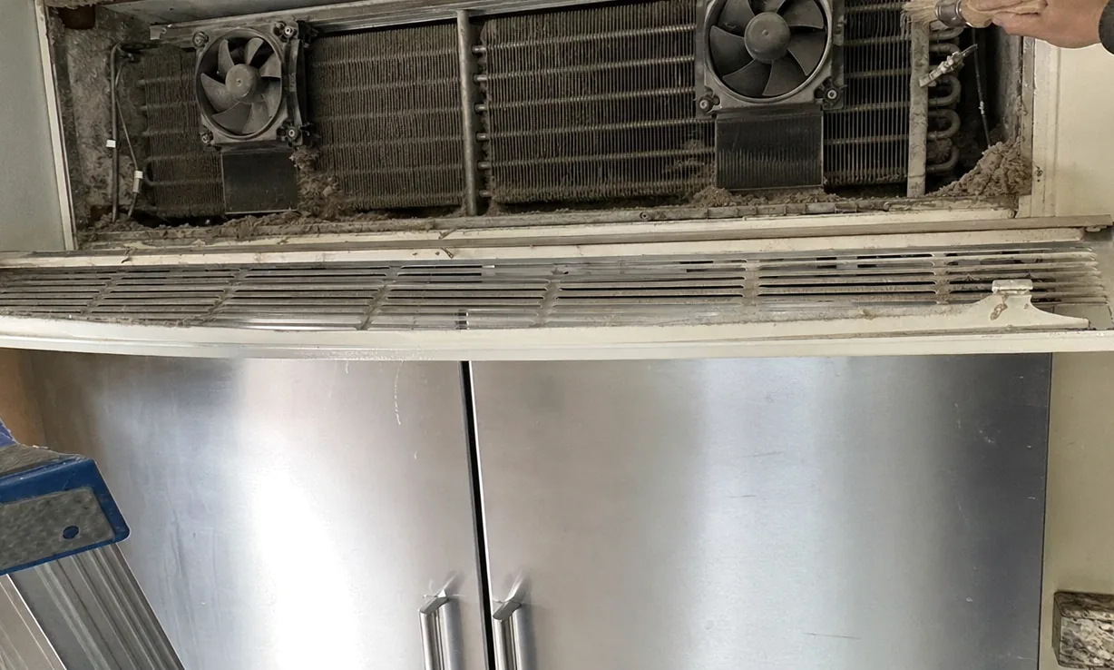 The louvred vent grille of a built-in refrigerator resting across the top of its stainless doors, felted dust along the slats and a granite worktop to the right.