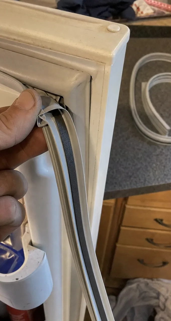 An older kitchen with a white appliance door standing open against timber drawer fronts, a hand holding the door gasket clear of the frame.