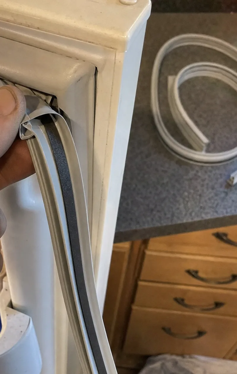 A hand peeling one corner of a refrigerator door gasket back from the door frame, the magnetic strip inside it visible against the white liner.